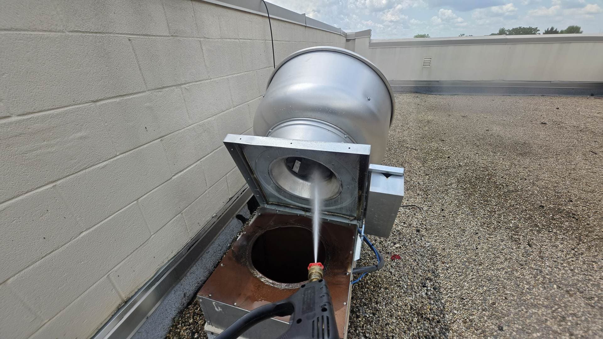 Rooftop upblast exhaust fan tipped back on its hinge while a technician pressure-washes down the grease duct through the open curb
