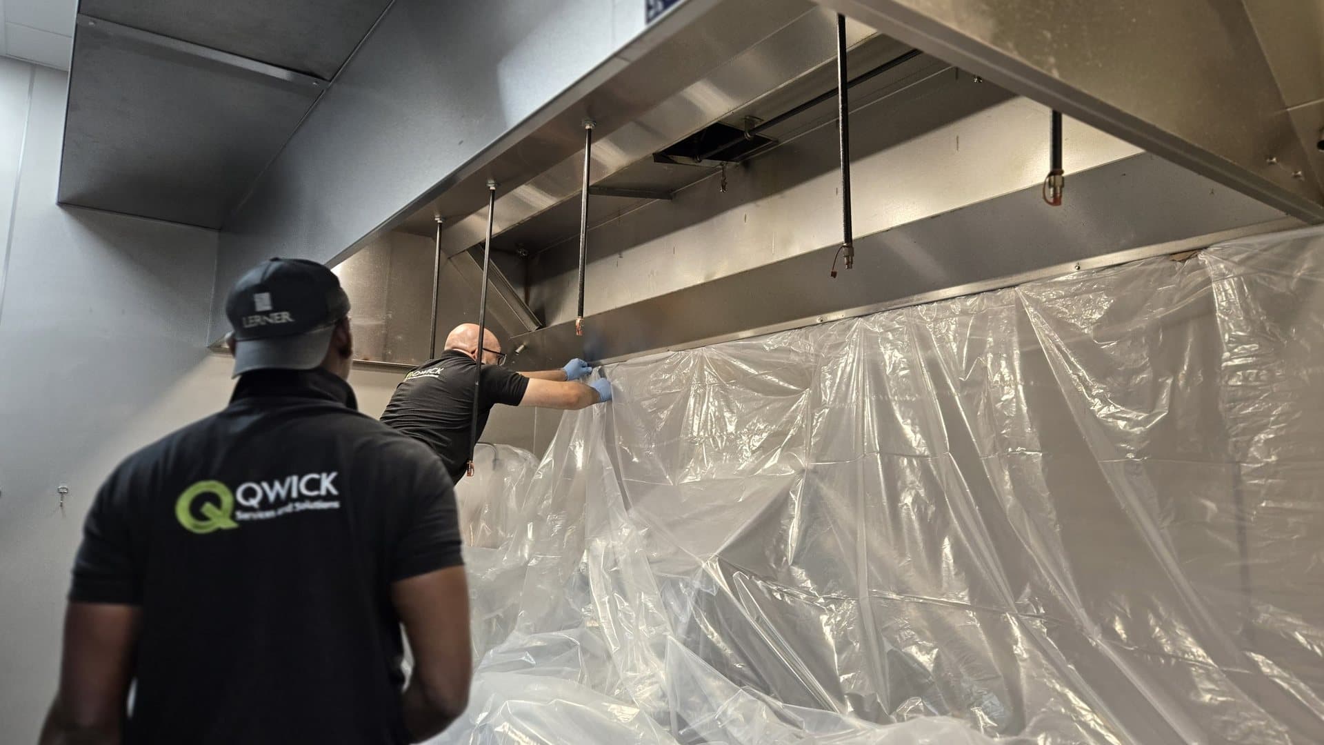 Two Qwick technicians taping clear plastic sheeting from a kitchen hood down over the cook line before a hood cleaning